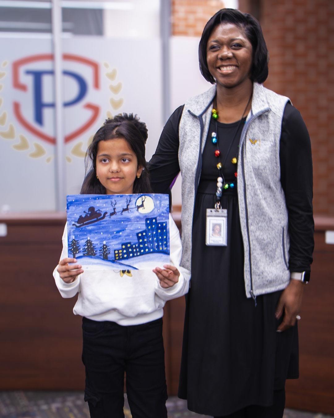 A school principal stands next to a student artist, both smiling, while holding or standing beside the student’s winter painting featuring Santa’s sleigh flying over a snowy nighttime scene.