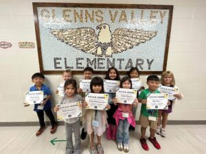 photo of ten first grade students standing in front of a tile mosaic of an eagle. Students are holding their Soaring eagle award certificate