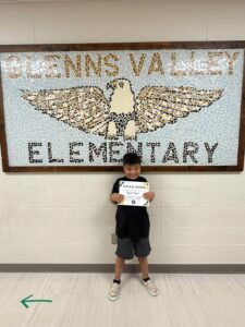 photo a third grade student standing in front of a tile mosaic of an eagle. Bash is holding his Soaring eagle certificate