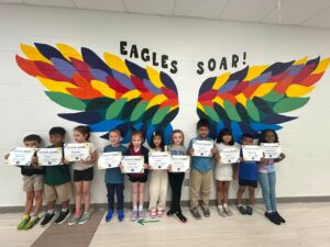 photo of eleven first grade students standing in front of the rainbow eagle wings. Students are holding their Soaring eagle certificate