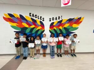 photo of nine second grade students standing in front of the rainbow eagle wings. Students are holding their Soaring eagle certificate