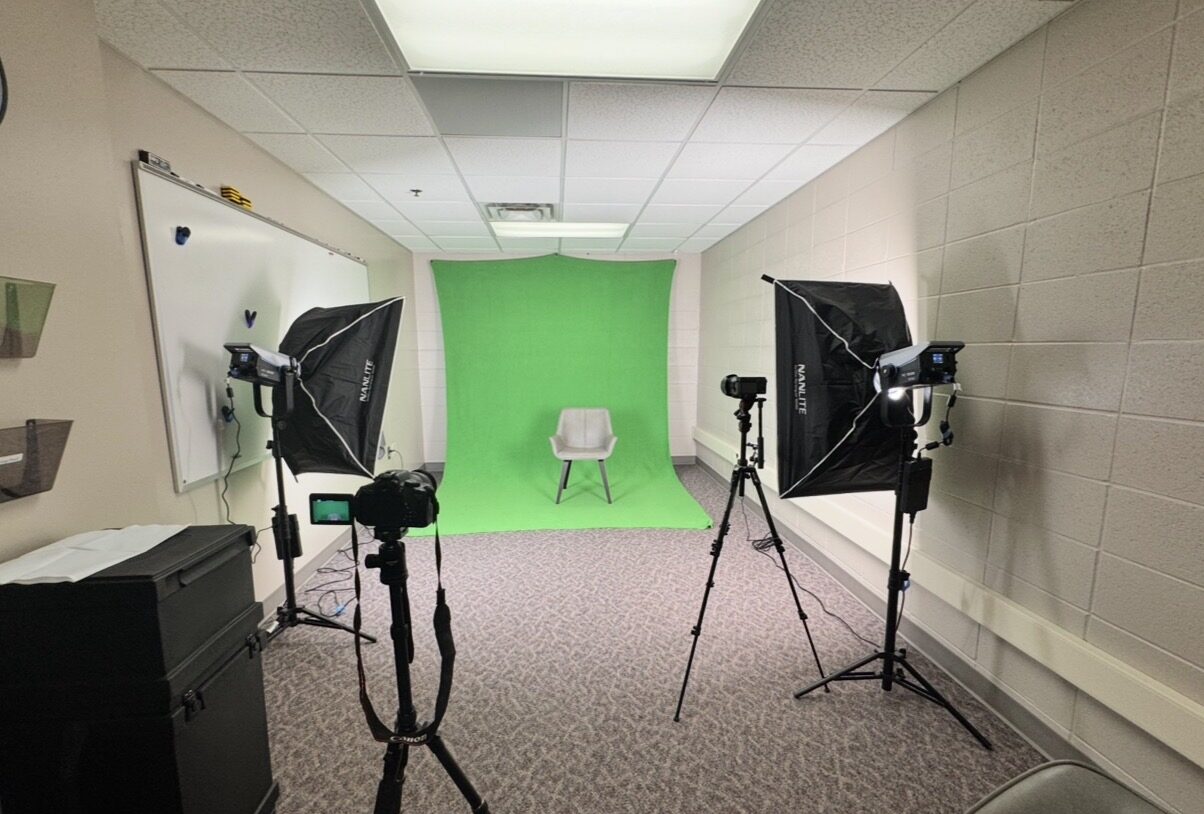Small indoor video studio setup with a green screen backdrop, a single chair centered in front, two softbox lights on stands, and a camera mounted on a tripod facing the chair.