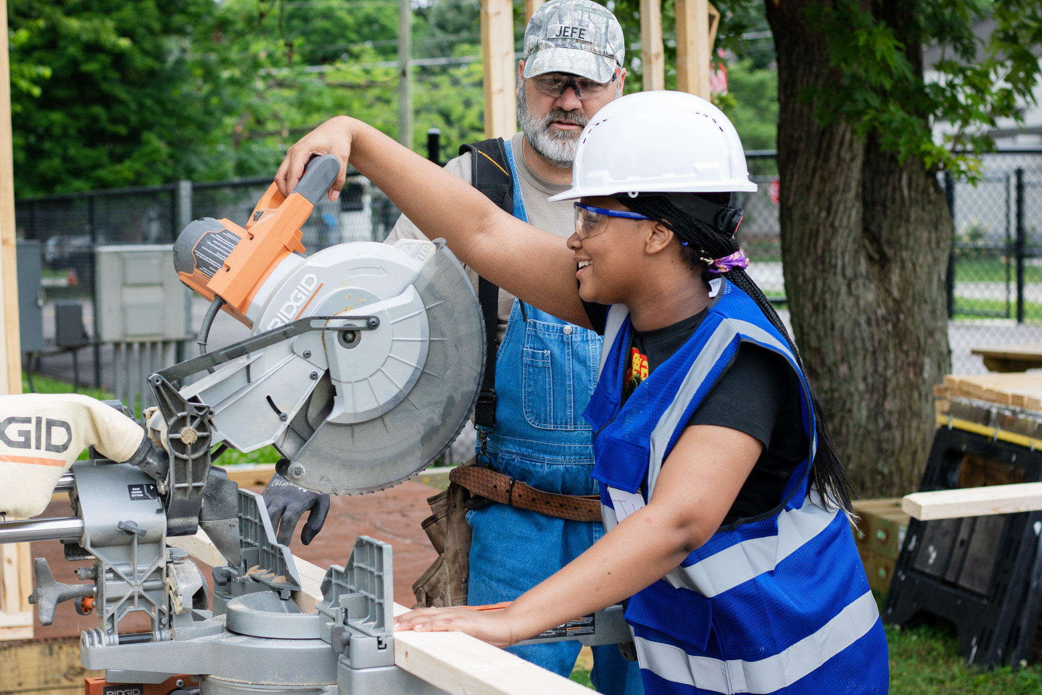 Student wearing a hard hat and safety vest uses a miter saw to cut wood while an adult instructor supervises and guides her hands at an outdoor construction site.