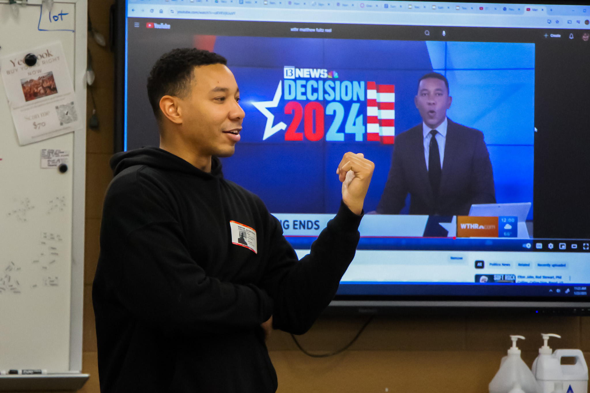 Mentor presents in front of a screen displaying a news broadcast about the 2024 election, gesturing while speaking to the class.