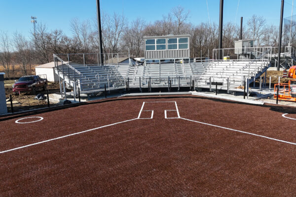 Ground-level view from home plate showing new synthetic turf infield, bleachers, press box, and dugouts at the newly constructed softball field.