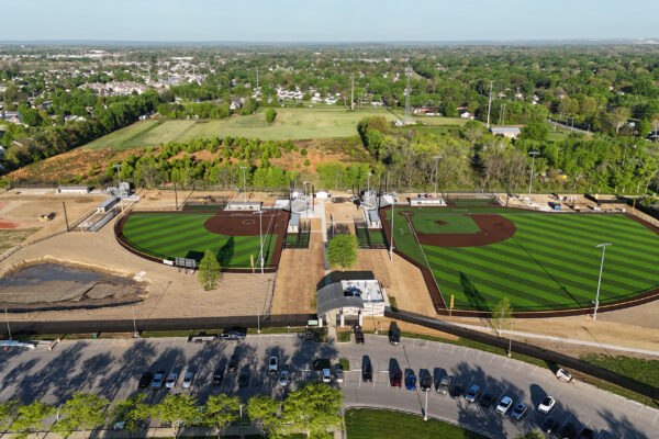 Aerial view of the full Holder Field complex showing two completed synthetic turf baseball and softball fields, surrounding walkways, parking lot, and neighboring landscape.