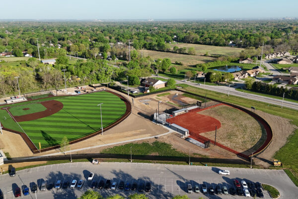 Aerial view of one completed synthetic turf field beside a second field still under construction, with surrounding roads, neighborhood, and open green space.