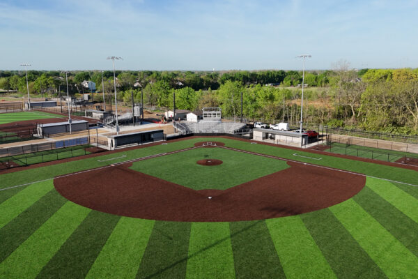 Aerial view of a newly completed baseball field with striped synthetic turf, brown infield, dugouts, bleachers, and adjacent softball field visible in the background.