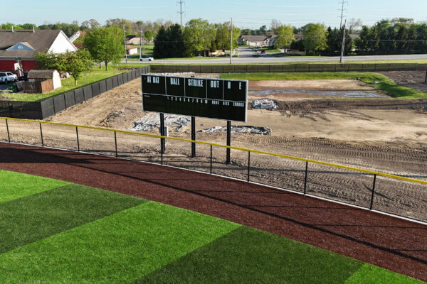 View of a new outfield scoreboard installed beyond the fence of a synthetic turf baseball field, with construction area visible behind it.