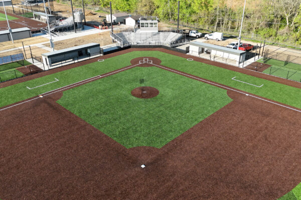 Aerial view of a newly constructed softball field featuring striped synthetic turf, brown infield, dugouts, backstop, bleachers, and press box behind home plate.