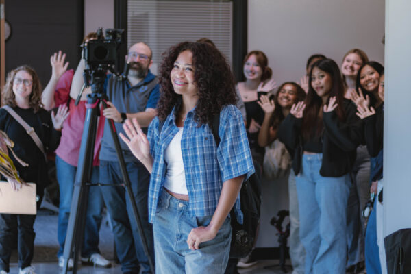 A Perry Township Schools student smiles and waves while walking through a learning space on the University of Indianapolis campus during filming for the Innovation & Entrepreneurship Program. Other students wave in the background as a videographer captures footage for the program's promotional video.
