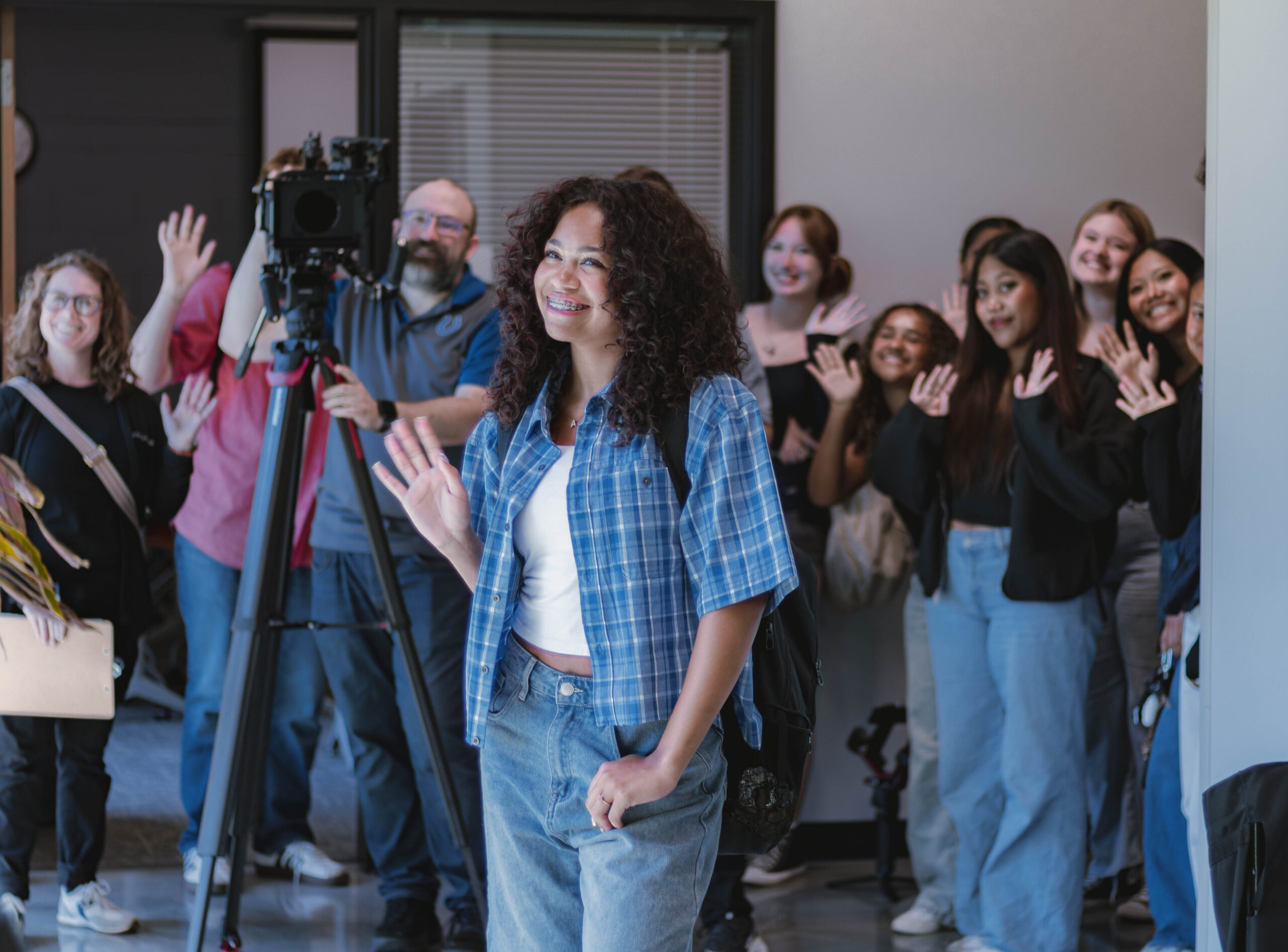 A Perry Township Schools student smiles and waves while walking through a learning space on the University of Indianapolis campus during filming for the Innovation & Entrepreneurship Program. Other students wave in the background as a videographer captures footage for the program's promotional video.