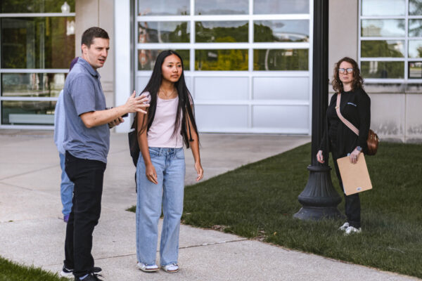 A student stands on a campus walkway while an adult gives directions during filming for Perry Township Schools' Innovation & Entrepreneurship Program. Another adult stands nearby holding a clipboard.