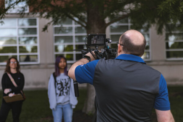 A student wearing a backpack walks along a campus sidewalk while a videographer records footage with a camera on a tripod. Another student stands near a building in the background.