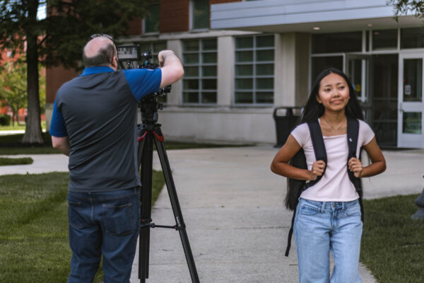A student wearing a backpack walks along a campus sidewalk while a videographer records footage with a camera on a tripod. Another student stands near a building in the background.