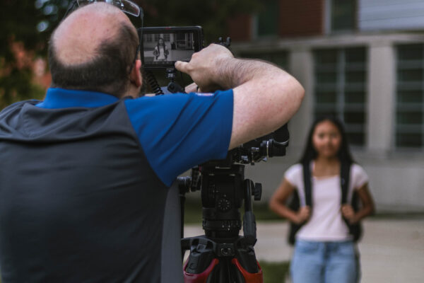 A student wearing a backpack walks along a campus sidewalk while a videographer records footage with a camera on a tripod. Another student stands near a building in the background.