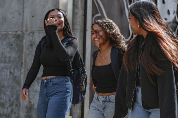 Three students walk side by side outdoors, smiling and talking while carrying backpacks on a college campus.