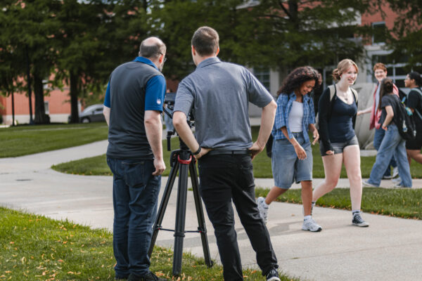 Members of the filming team stand beside a camera while students walk across the University of Indianapolis campus during production for Perry Township Schools' Innovation & Entrepreneurship Program.