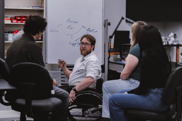 A business professional using a wheelchair leads a discussion with students gathered around a whiteboard during a collaborative learning session for Perry Township Schools' Innovation & Entrepreneurship Program.