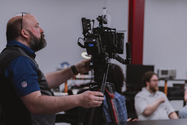 A videographer operates a camera while capturing footage of students and a business professional during filming for the Innovation & Entrepreneurship Program.